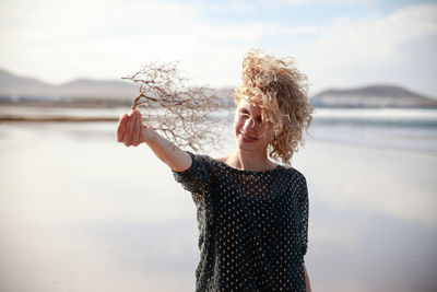 Smiling woman holding dry plant at beach against sky