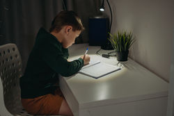 Caucasian boy doing homework at home in dark room by the light of lamp.