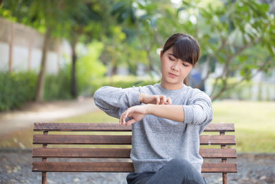 Young woman scratching hand while sitting on bench at park