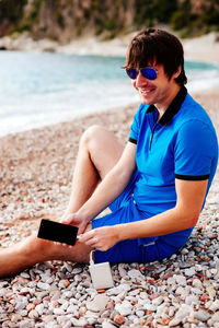 Smiling man using mobile phone while sitting on pebbles at beach