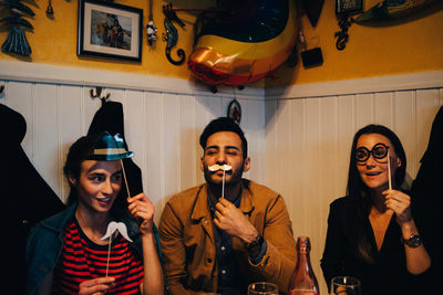 Cheerful young friends holding props while sitting at restaurant during dinner party