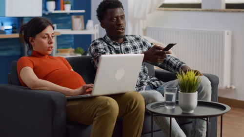 Pregnant woman using laptop while husband watching movie at home