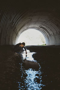 Side view of boy looking into water while crouching in tunnel