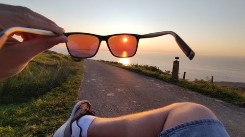 Low section of person wearing sunglasses against sky during sunset