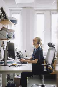 Side view of happy female healthcare worker sitting and using computer at desk in clinic