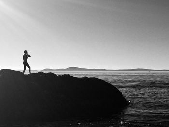 Silhouette man standing on rock by sea against clear sky