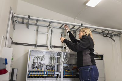 Female electrician fixing cable wires on ladder in meter room at industry