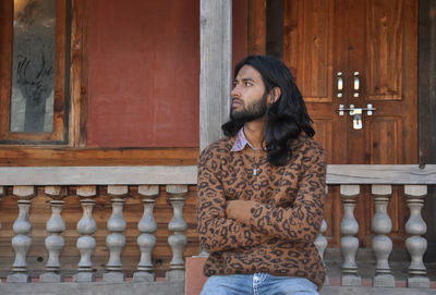 A long haired young guy looking sideways while sitting in old temple posing with crossed arms 