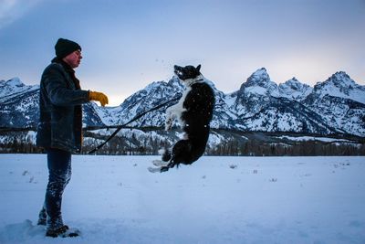 Side view of man playing with dog by mountains on snowy field