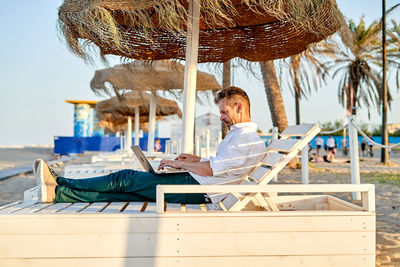 Side view of focused mature male manager in smart casual clothes working on laptop while sitting on lounger on summer day on beach