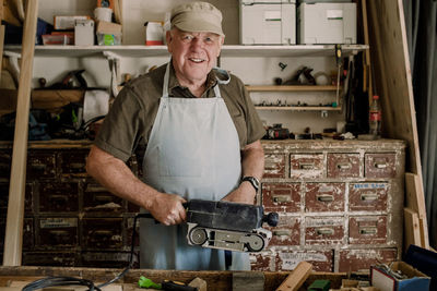 Portrait of confident smiling senior male owner holding power tool at workshop