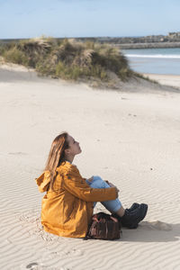 Young woman hugging knees while sitting at beach during sunny day