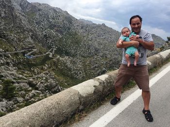 Portrait of man carrying baby while standing on road against mountain