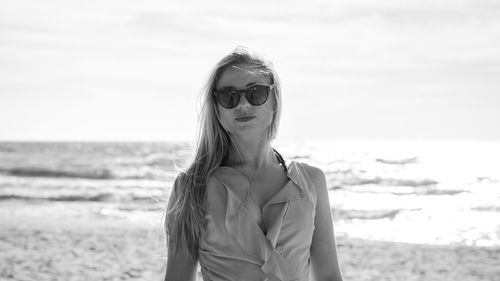 Portrait of mid adult woman wearing sunglasses while standing at beach against sky