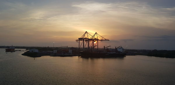 Cranes at harbor against sky during sunset