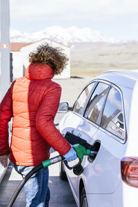 Side view of serious male with curly hair in outwear and latex gloves standing while refueling car at gas station in sunny day