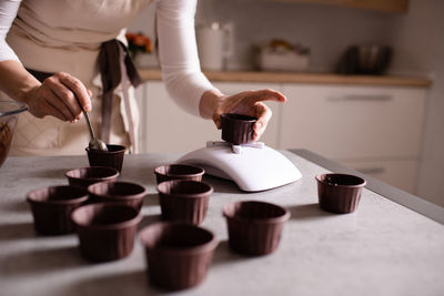 Woman making chocolate cupcakes weigh forms on kitchen scale on table close up.