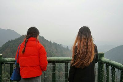 Rear view of women standing by railing while looking at mountains in foggy weather