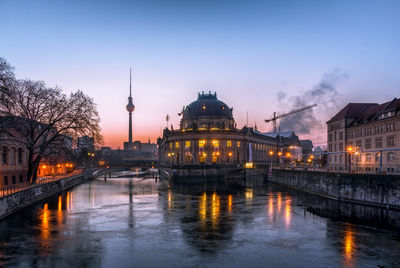 Illuminated buildings by river at dusk