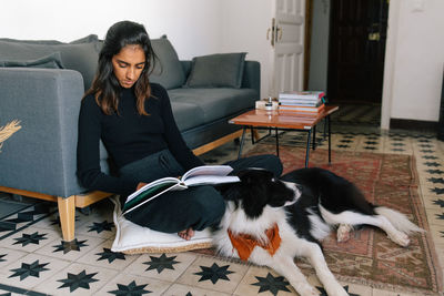 Thoughtful indian female reading interesting story in book and sitting on floor with fluffy friendly border collie dog