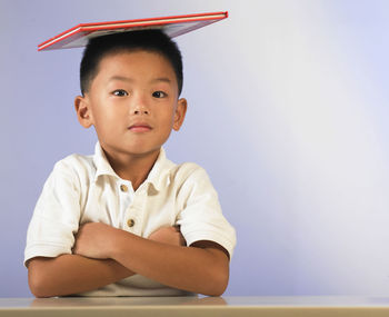 Portrait of boy balancing book while sitting with arms crossed at table against wall