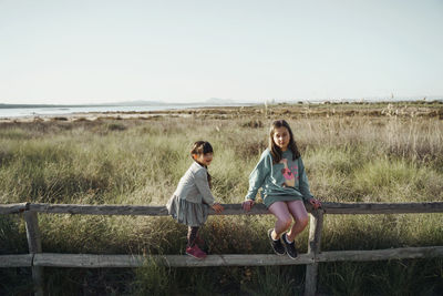 Full length of girls  sitting on woden fence among field