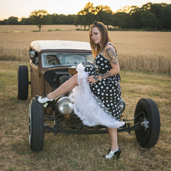 Full length side view portrait of tattooed young woman standing by vintage vehicle on field
