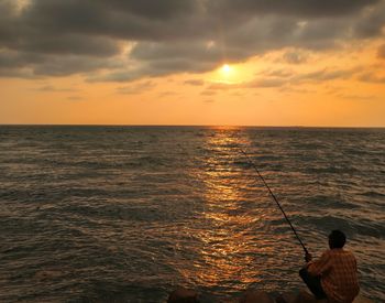 Man fishing in sea against sky during sunset