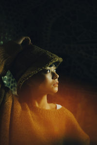 Young woman wearing hood while looking away at illuminated restaurant