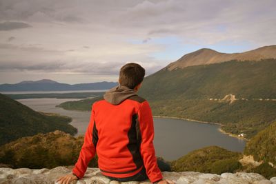 Rear view of man sitting on cliff lake and mountains against cloudy sky