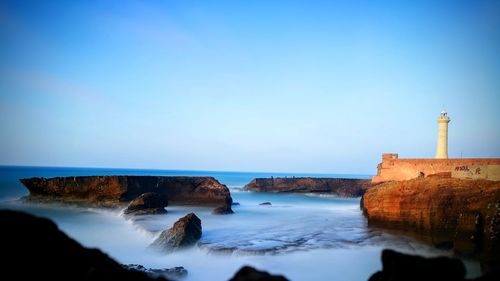 Lighthouse amidst sea and buildings against clear blue sky