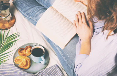 High angle view of woman reading book while having breakfast on bed