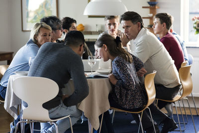 Girl and man reading magazine while sitting with family and friends at dining table