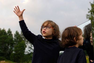 Boy standing with hand raised while brother looking at mirror in park against sky