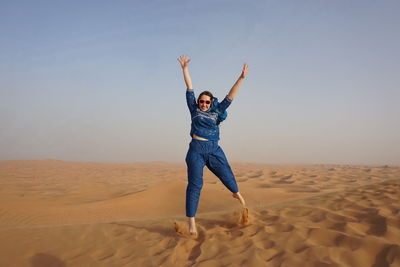 Full length of excited woman jumping at desert against clear sky