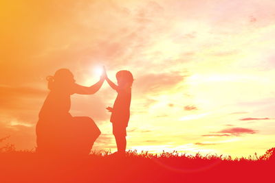 Silhouette mother giving high-five to daughter on field against sky during sunset