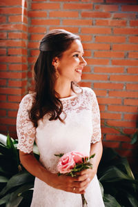 Smiling woman holding flowers while standing against brick wall