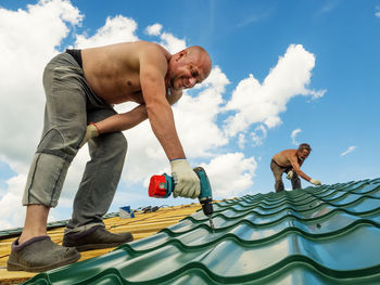 Shirtless men working on roof against sky