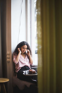 Young woman looking away while sitting on window at home