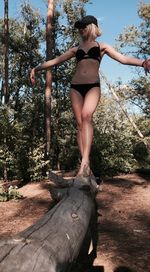 Young woman wearing bikini walking on log against sky in forest