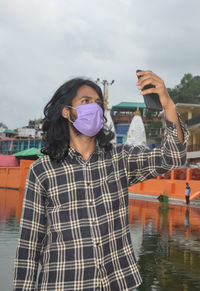 A south asian young man with long hair taking selfie with wearing face mask while standing outside 