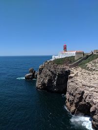 Lighthouse on rocks by sea against clear blue sky