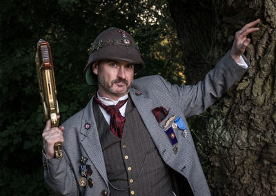 Portrait of man holding gun while standing by tree