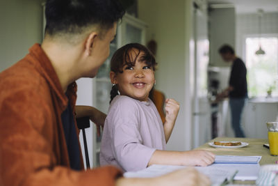 Cheerful girl holding fist and looking at father while doing homework at home