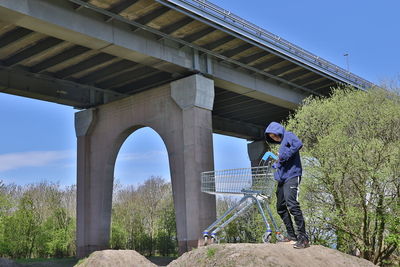 Full length of boy with shopping cart standing on rock against bridge during sunny day