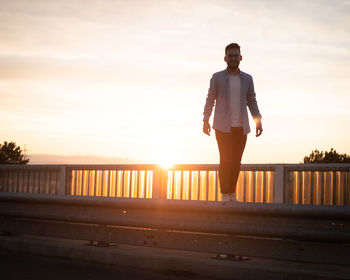 Portrait of man standing on crash barrier against sky during sunset