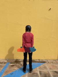 Rear view of teenager holding skateboard facing a yellow wall