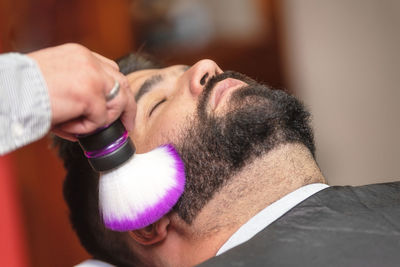 Cropped hand of barber using shaving brush on customer beard in salon