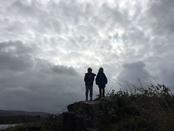 Siblings standing on field against cloudy sky