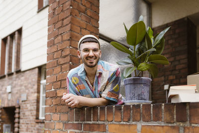Portrait of smiling man leaning by potted plant on brick wall
